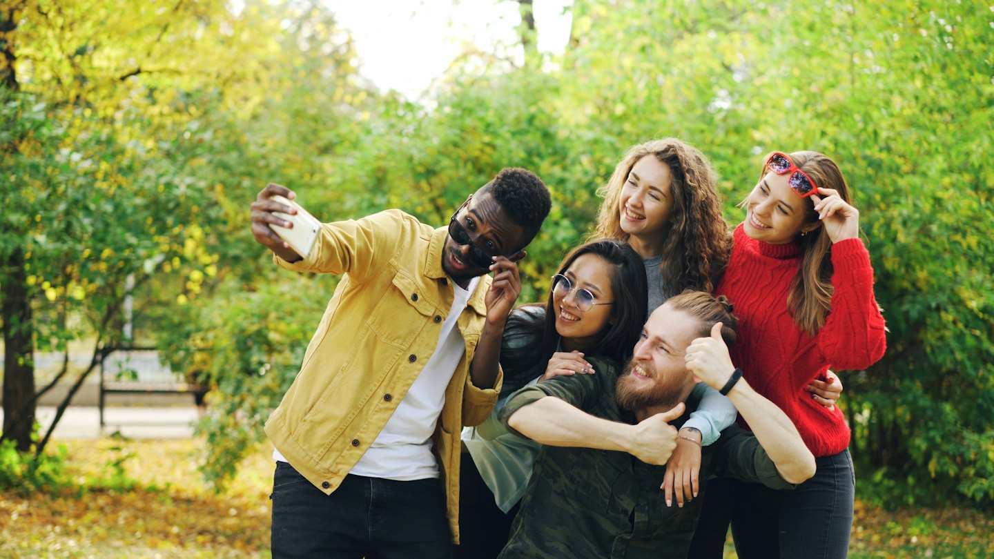 Five young friends smiling while taking a selfie in a park.
